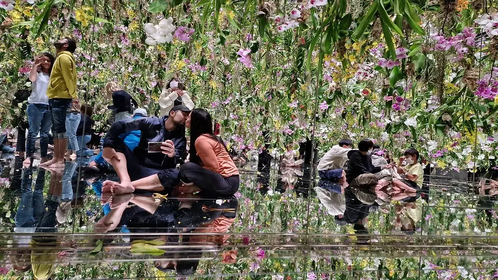 People sit and walk on a mirrored floor beneath hanging flowers. A couple kisses, others admire the vibrant floral display, creating a surreal scene.