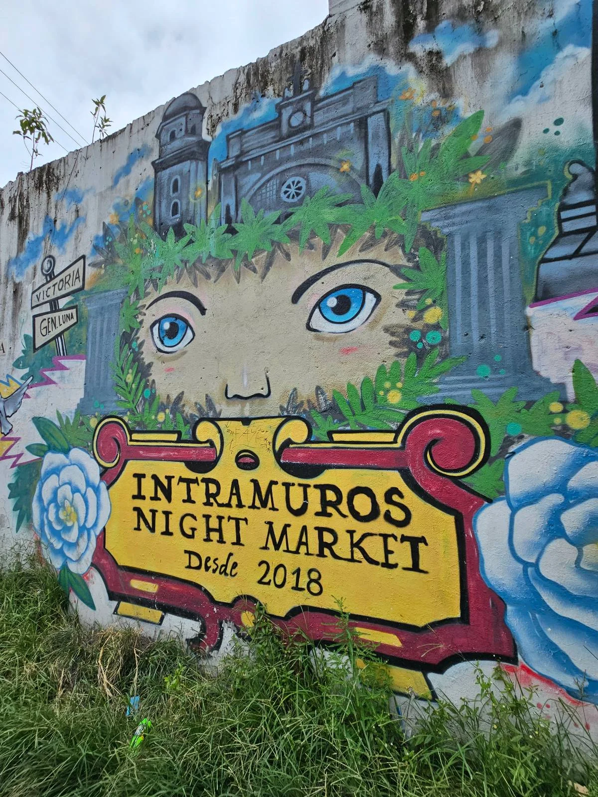 Colorful mural on a concrete wall featuring a pair of blue eyes peeking through greenery with historical buildings, blue flowers, and a yellow-red sign that reads "Intramuros Night Market Dec 2018.