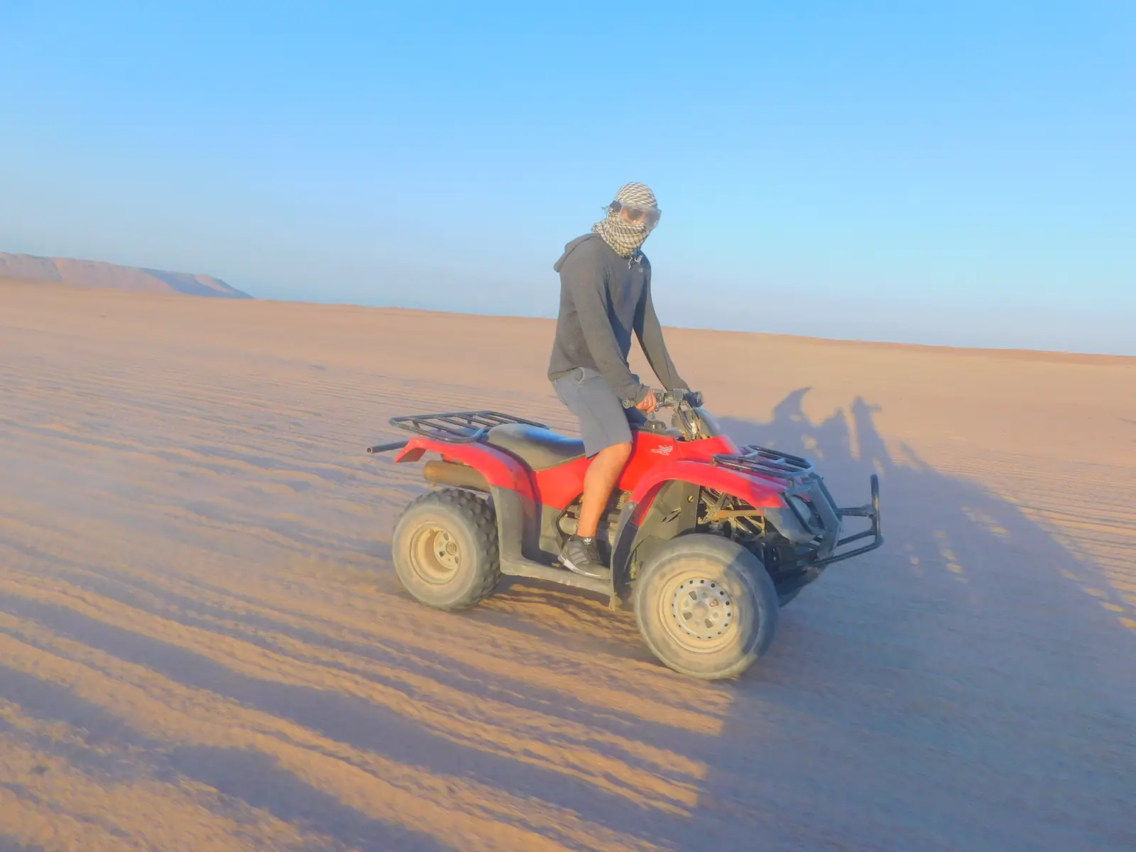 A person wearing a helmet is riding a red ATV across a vast, sandy desert under a clear blue sky. Long shadows are cast on the sand, and the terrain is flat and expansive.