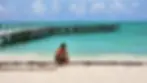 Man crouching on a white sand beach front close to the sea with a long wooden dock in the background at Passion Island, Quintana Roo, Mexico