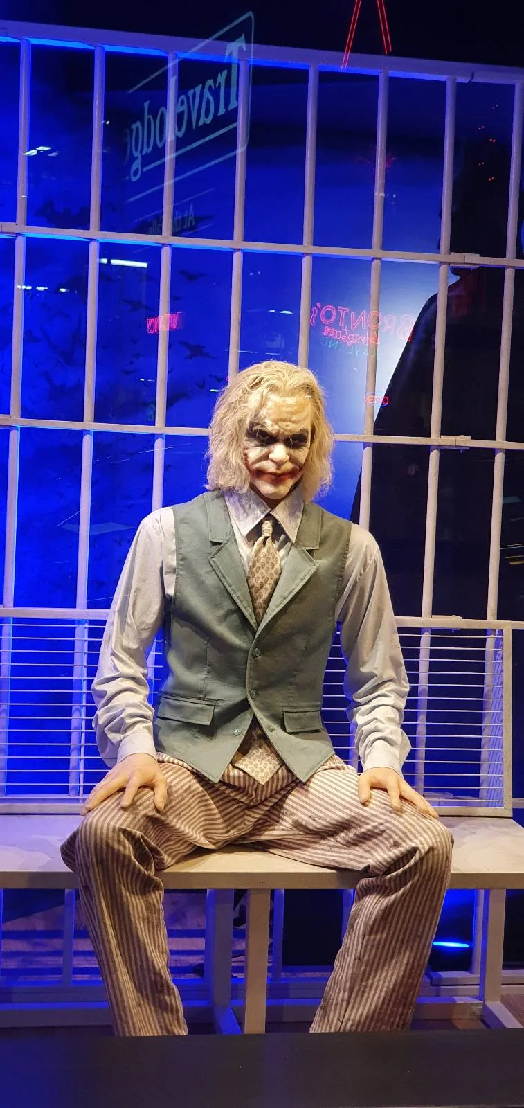 A person dressed as the Joker with white face makeup, red lips, and green hair sits on a bench in front of large jail bars, wearing a vest, shirt, and tie. Blue lighting fills the background.