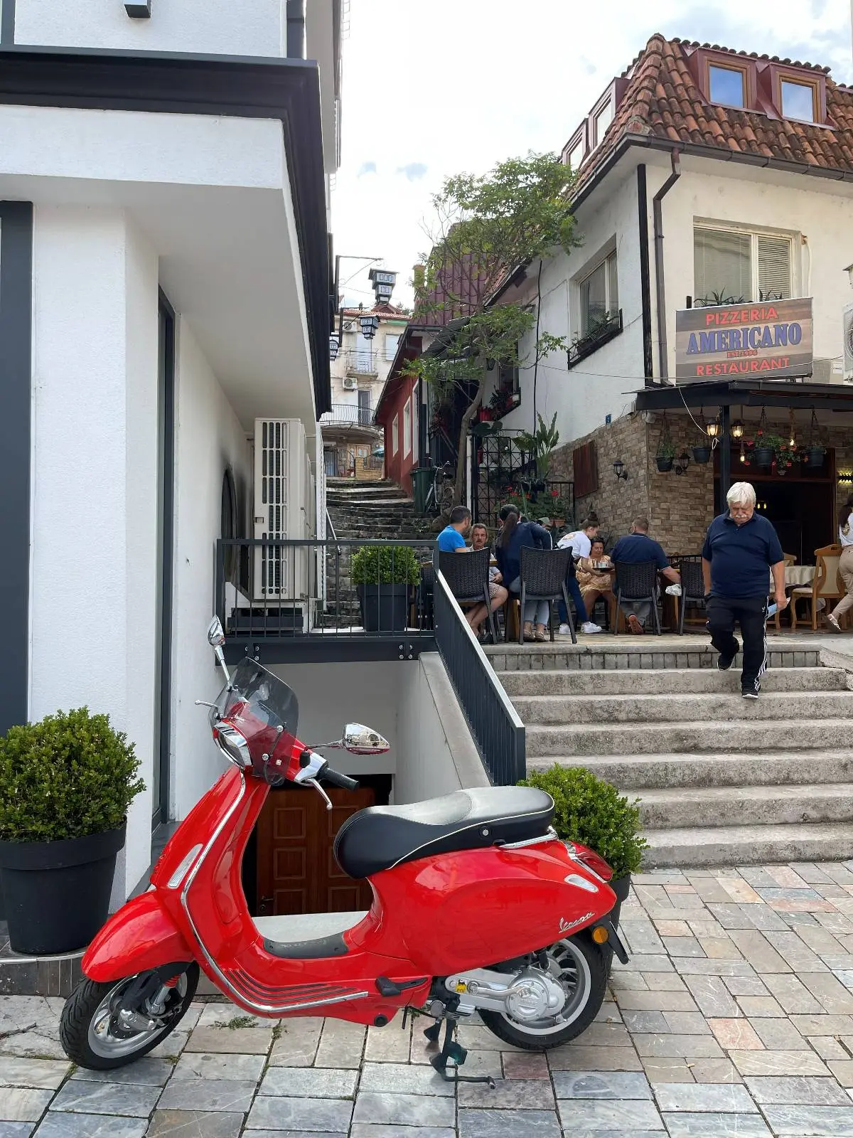 Red scooter parked on a cobblestone path beside a white building with potted plants. Stairs lead to an outdoor cafe where people are seated and walking. A rustic building with a sign and Mediterranean-style architecture appears in the background.