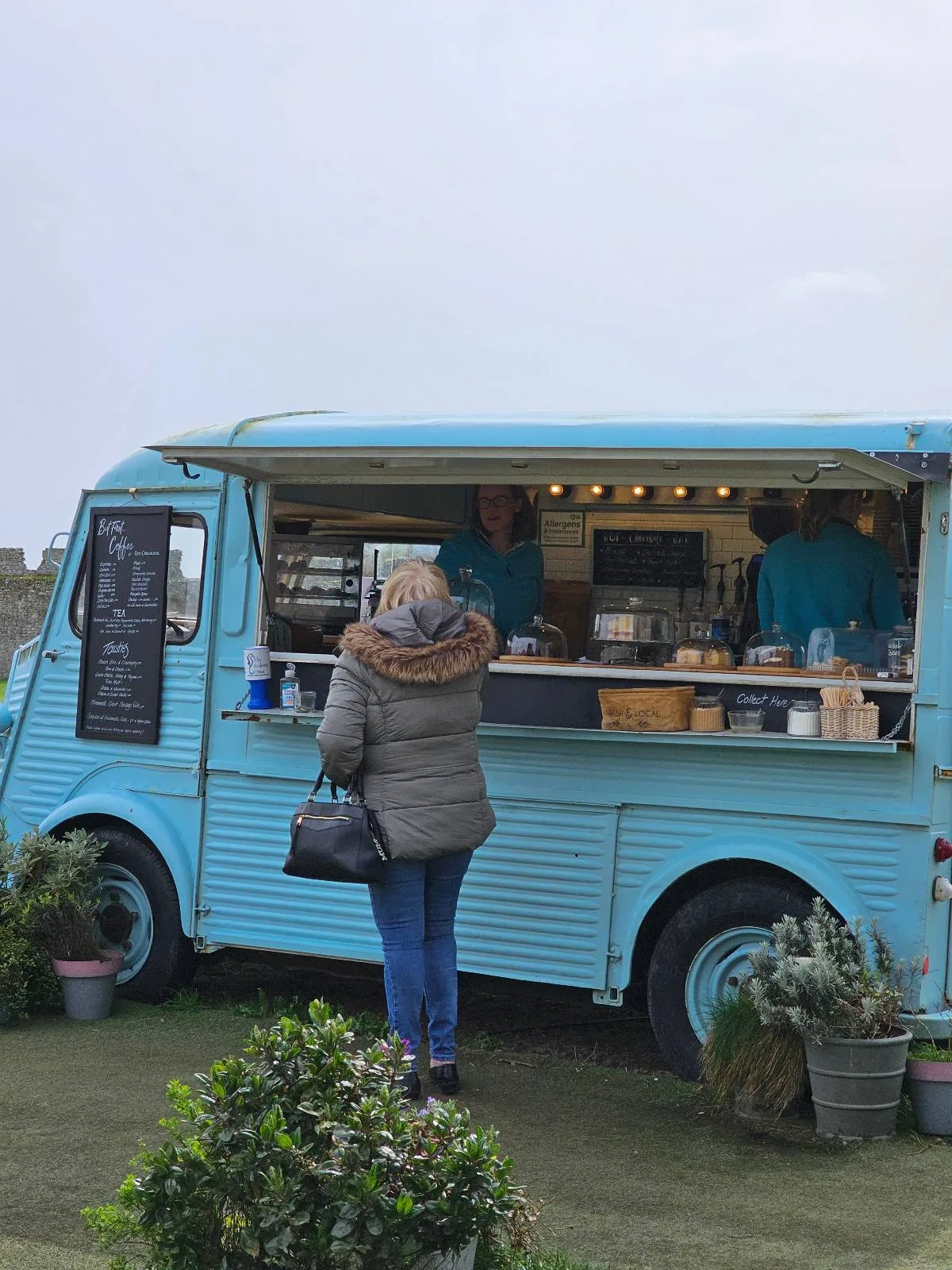 A customer ordering from a blue food truck with two people inside, parked outdoors with plants nearby.
