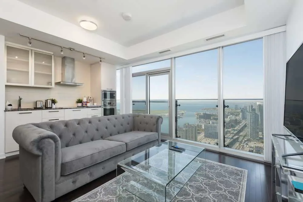Modern living room with a gray tufted sofa, glass coffee table, and a large TV. The area opens to a sleek kitchen with bar stools. Floor-to-ceiling windows offer a view of a city skyline and a body of water beyond.