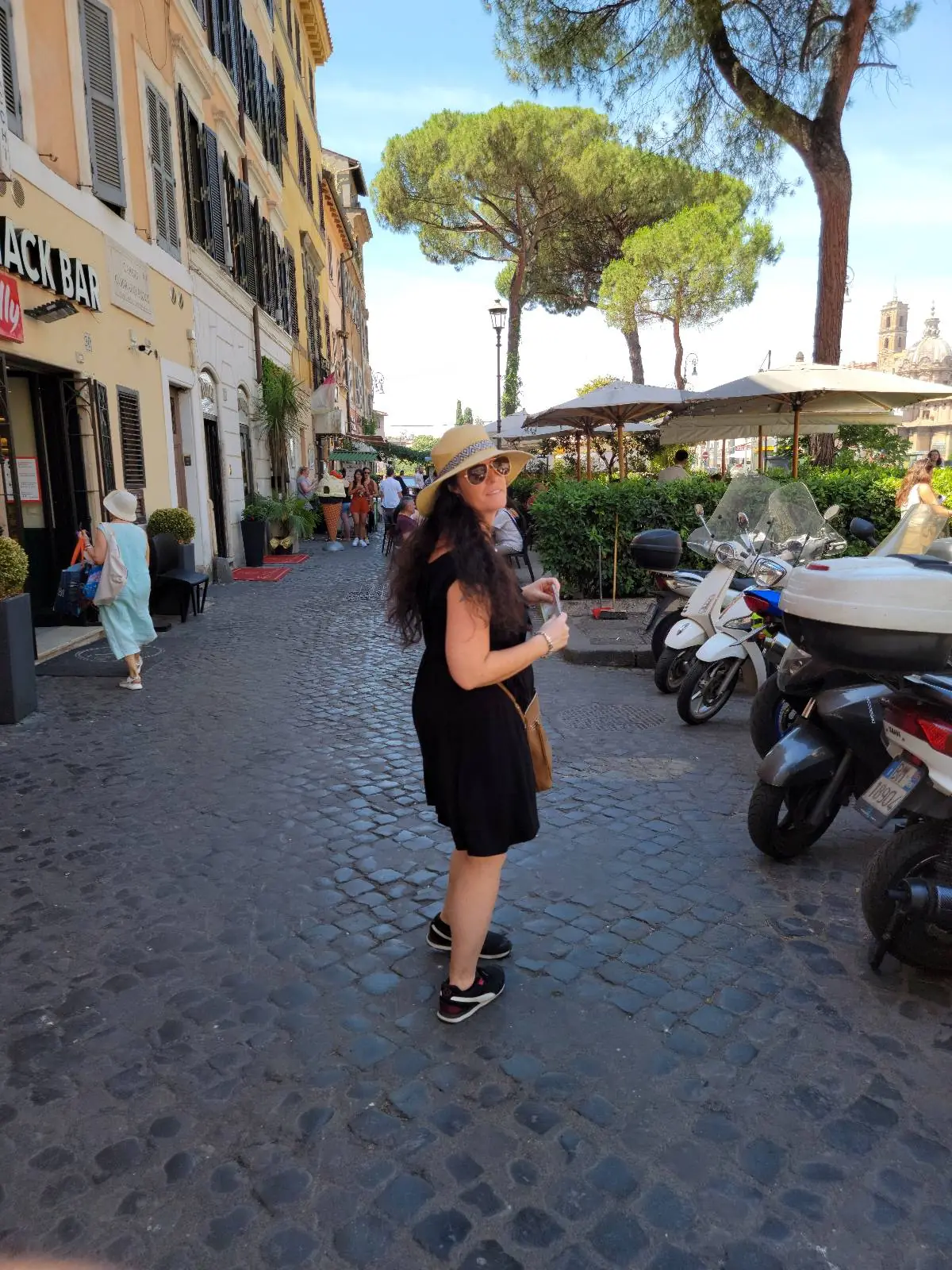 A woman in a black dress and straw hat stands on a cobblestone street lined with cafes, scooters, and trees, smiling back at the camera on a sunny day.