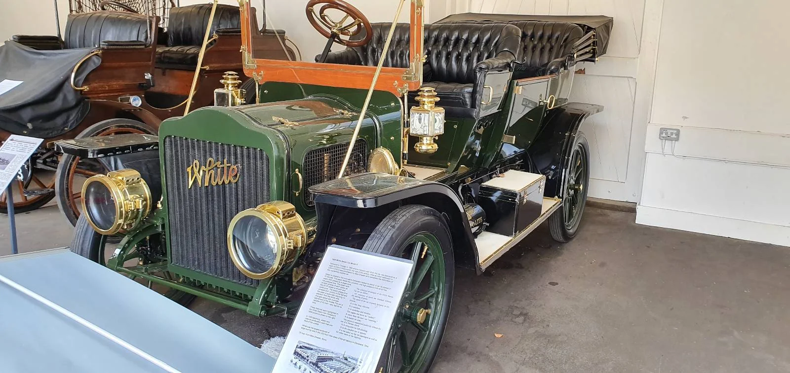 A vintage green car with a gold emblem on the front, displayed in a museum setting. It features brass headlights, a wooden steering wheel, and a black leather interior. Informational plaques are placed nearby.