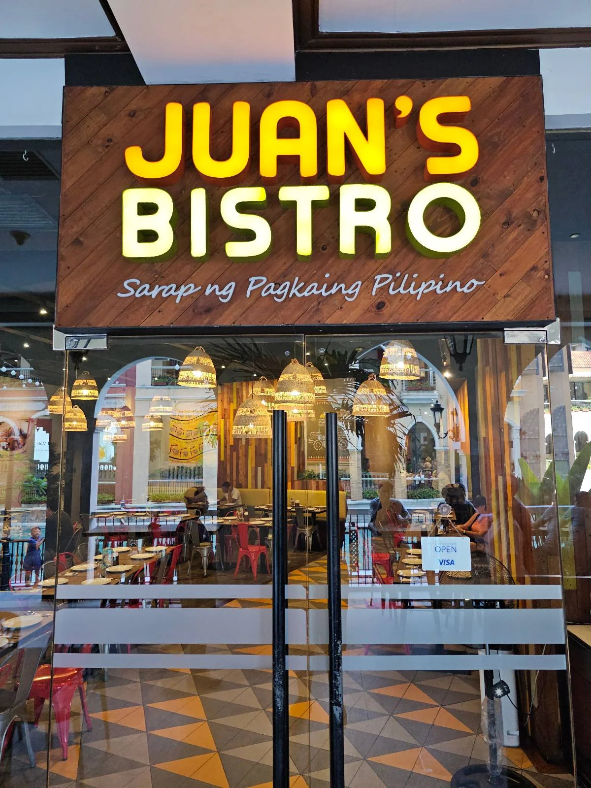 A restaurant entrance with a sign reading "JUAN'S Bistro, Home of Palabok Piyapeno." The interior has yellow hanging lights, red chairs, and several customers dining inside.