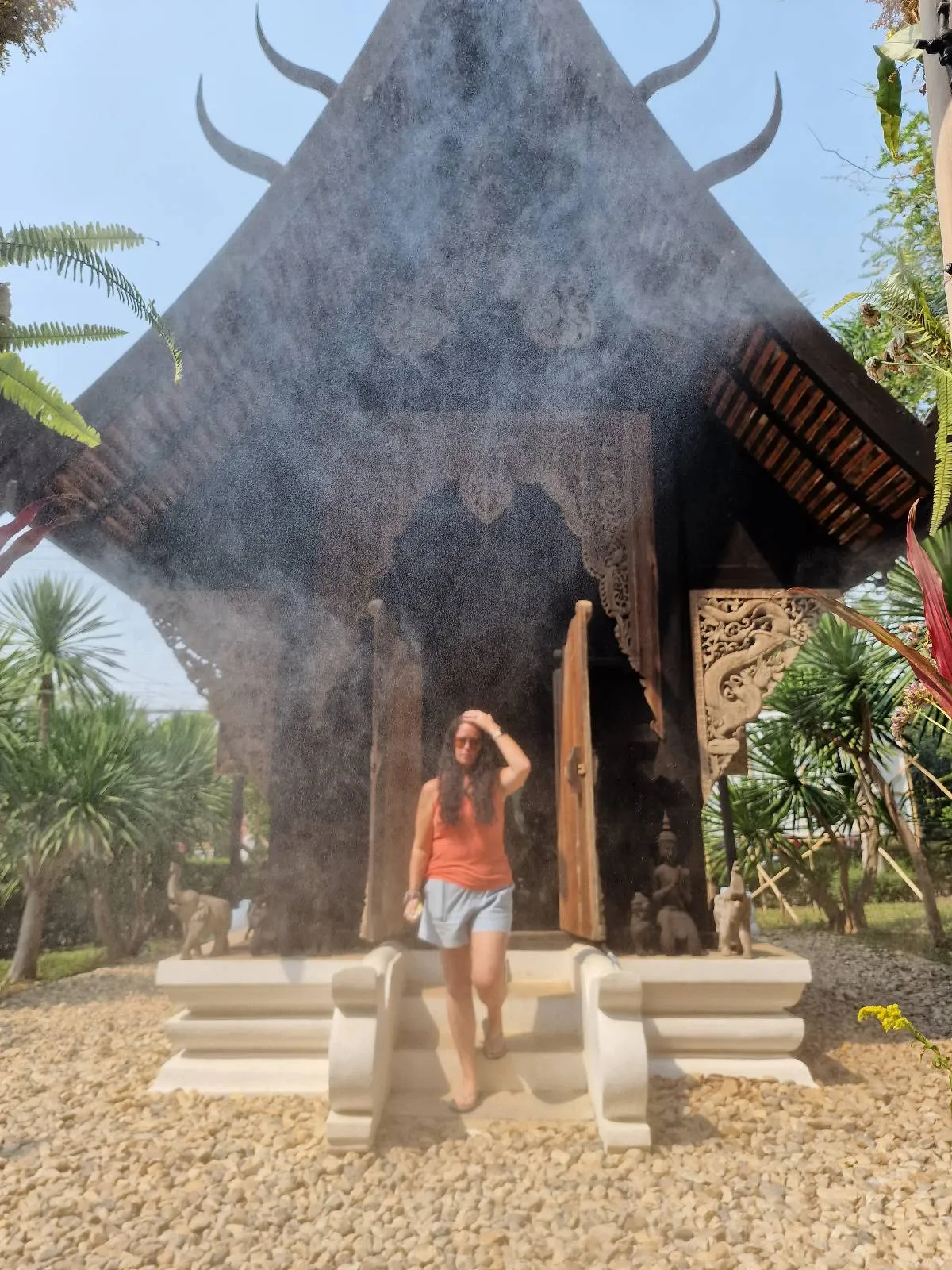 A person stands in front of a small, ornate building with a pointed roof, surrounded by greenery and smoke or mist in the air.