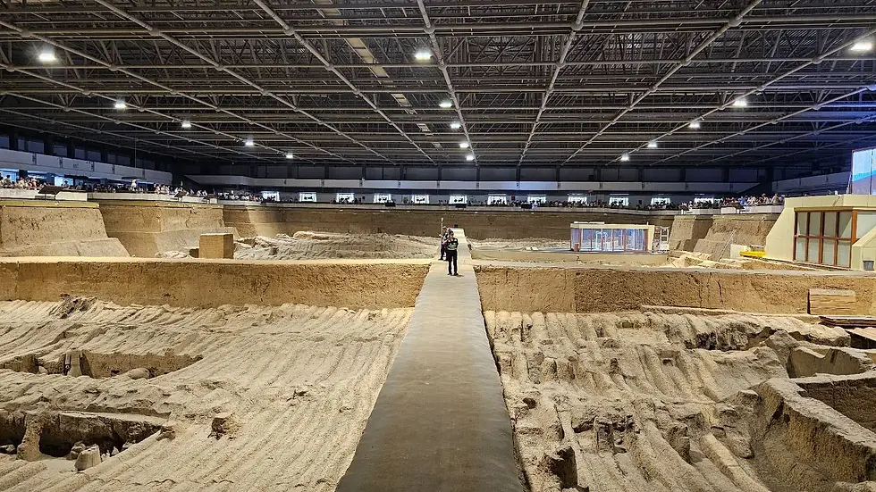 Large indoor archaeological site with sandy trenches and a metal ceiling. People observe from a distance. Quiet, museum-like atmosphere.