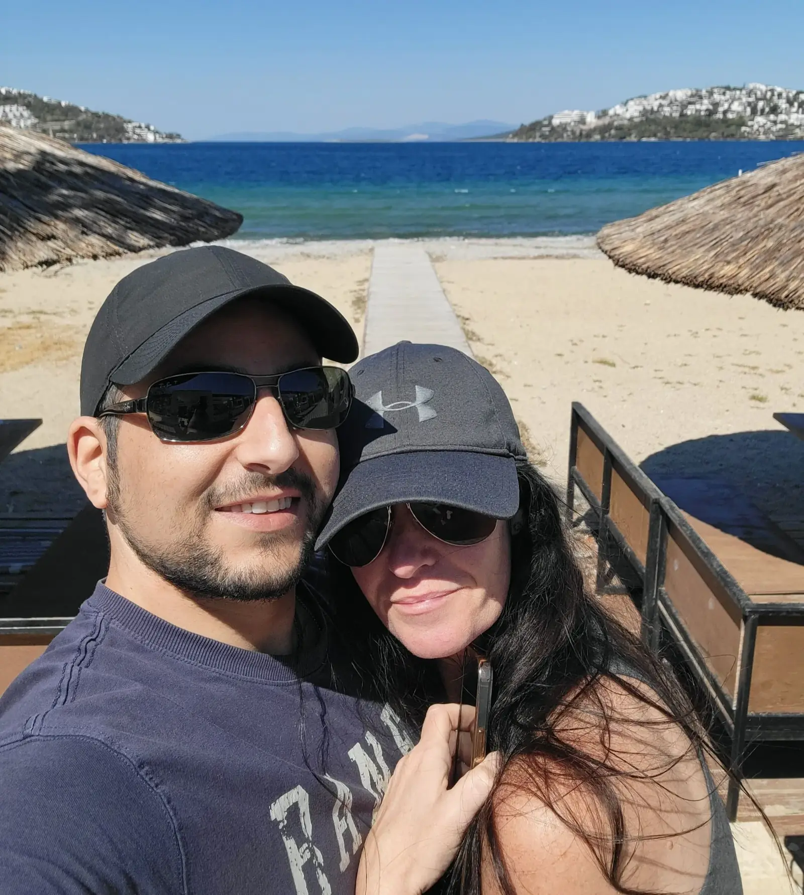 A couple wearing sunglasses and hats poses for a selfie on a sandy beach with clear blue water and hills in the background, under a sunny sky.