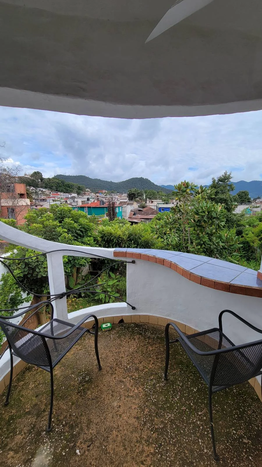 View from a curved balcony overlooking a small town with hills in the background. The balcony has two black metal chairs and a low wall. The sky is cloudy, and lush greenery surrounds the area.