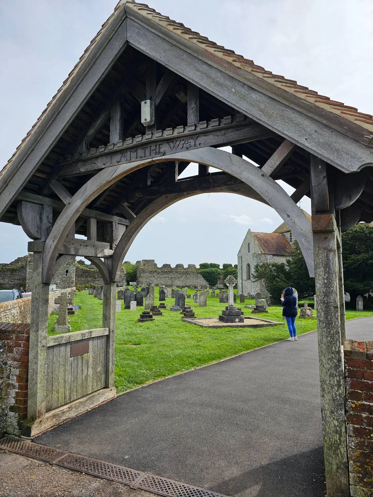 Wooden lychgate entrance to an old cemetery with gravestones and a church in the background, a person in blue walking under the arch.
