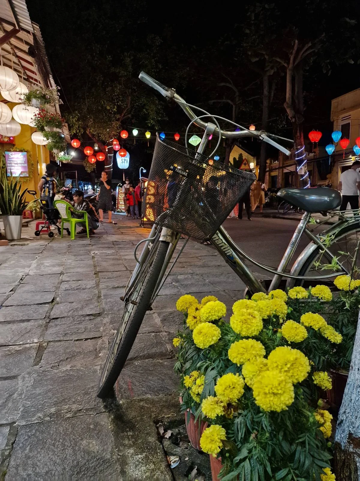 A vintage bicycle leans against a pot of bright yellow flowers on a cobblestone street at night, with colorful lanterns and people in the background.