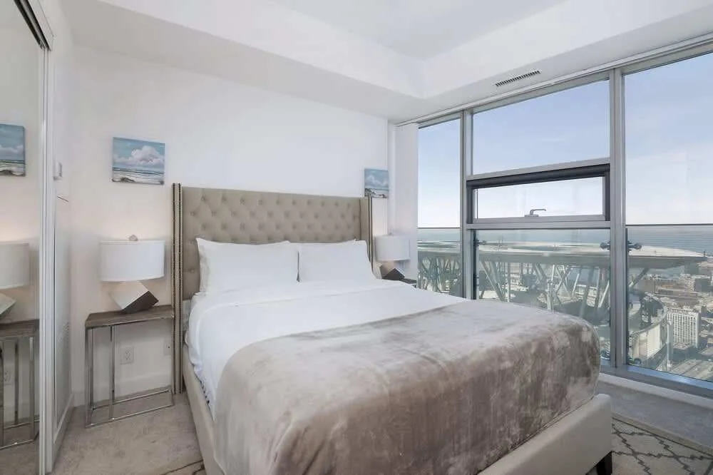 Bright bedroom with a tufted bed, white bedding, and gray throw. Large windows show a cityscape and ocean view. Soft, calming mood.