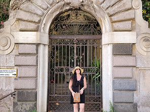 A woman in a black dress, sandals, and a hat stands before an ornate arched iron gate set in a stone wall with vines above it—capturing the charm of Rome on a layover. Cobbled pavement stretches underfoot.