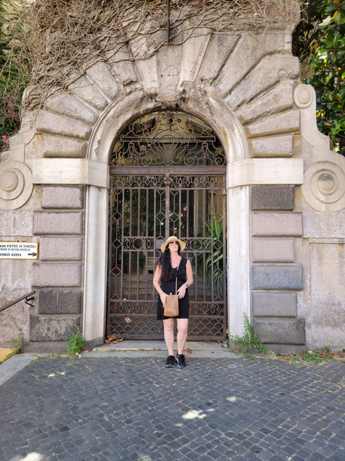 A person in a black dress stands in front of an ornate stone archway with a metal gate, surrounded by greenery and cobblestone pavement.