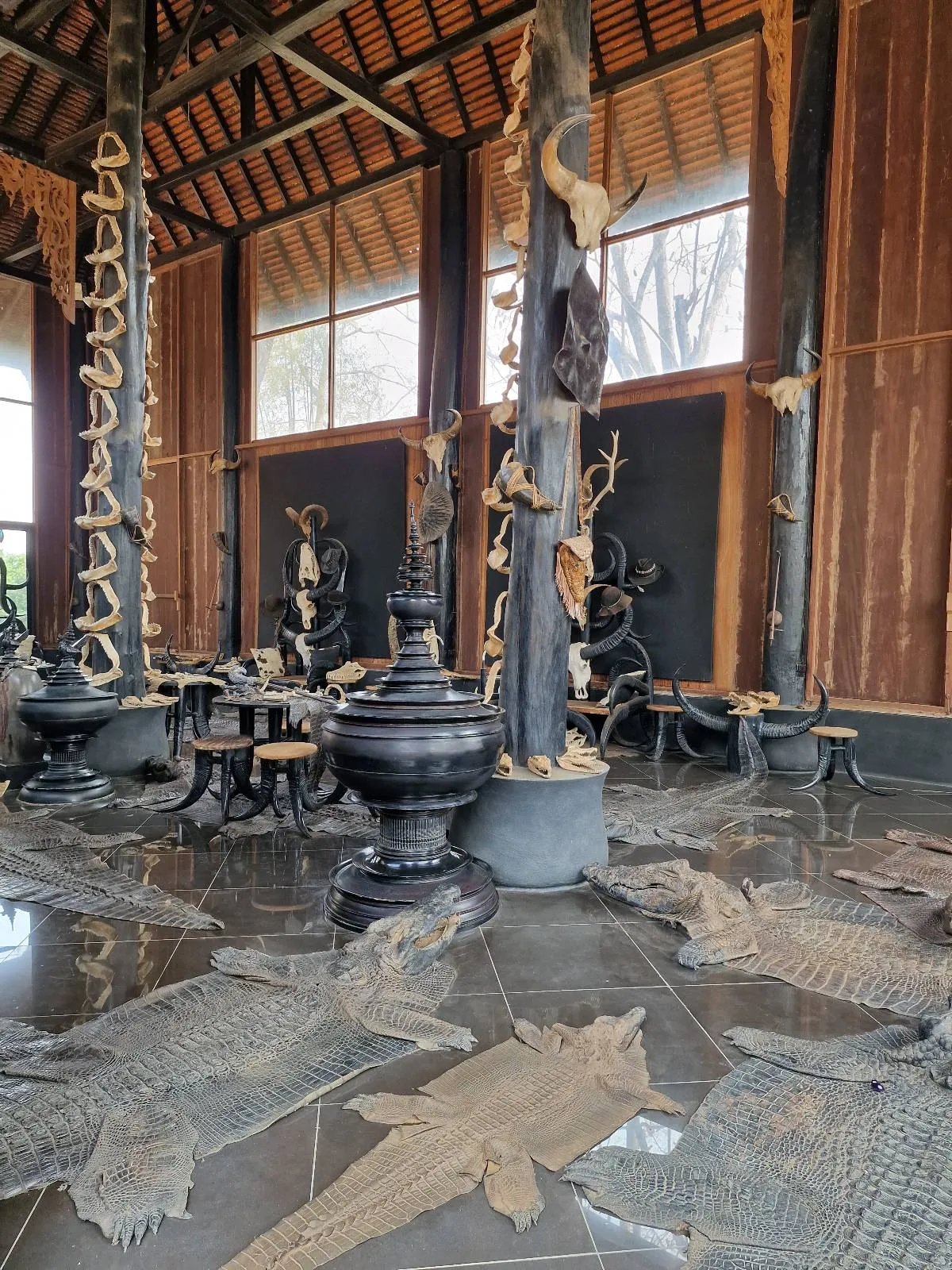Interior of a building with wooden pillars and intricate sculptures. The floor is decorated with woven mats resembling fish. Large windows allow natural light into the space.