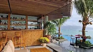 A modern rooftop bar with a bamboo-textured counter, wooden seating, and a view of an infinity pool overlooking the ocean and misty mountains.