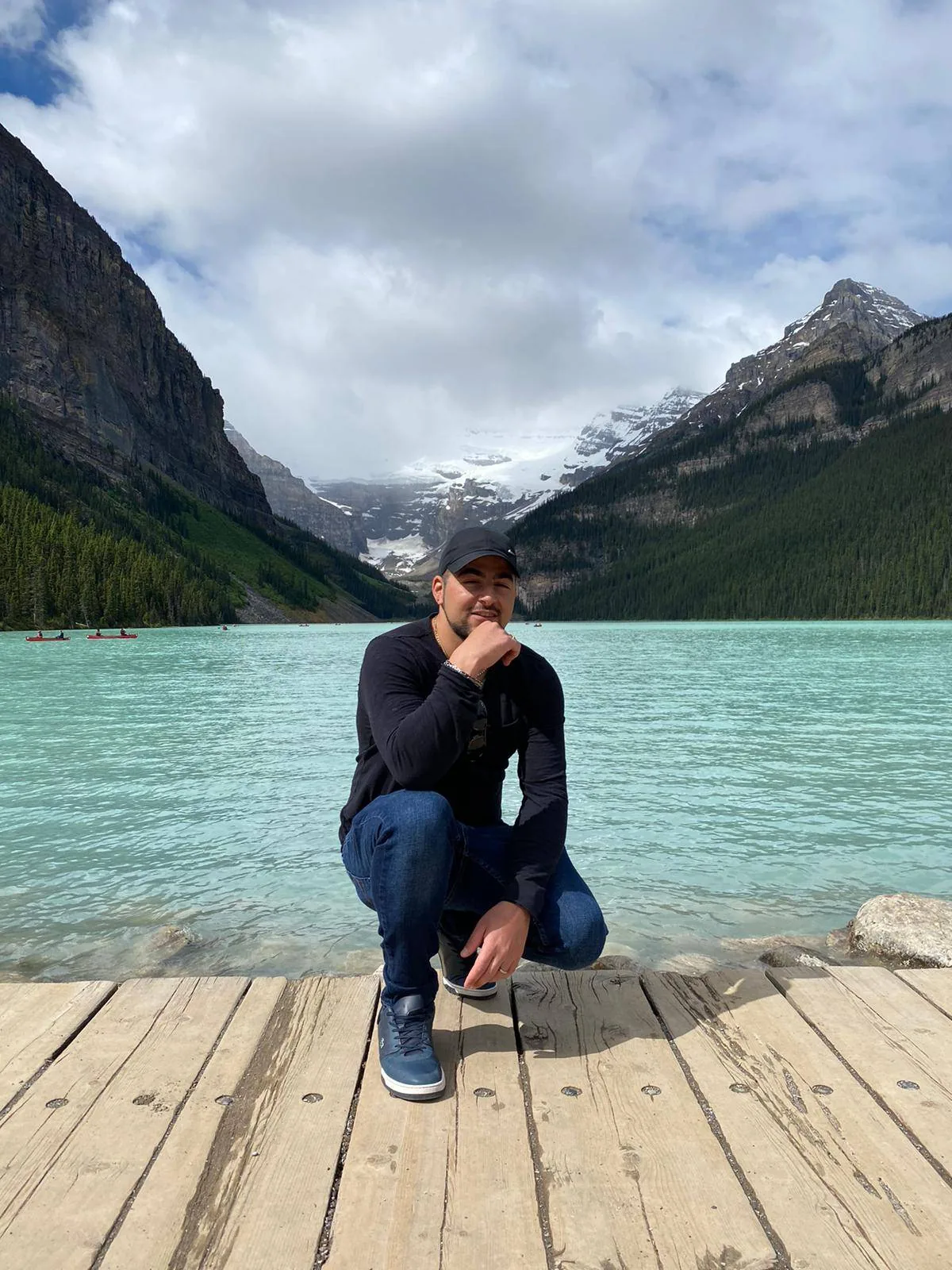 A person in a black sweater, jeans, and cap crouches on a wooden dock by a turquoise lake, surrounded by mountains and cloudy skies.