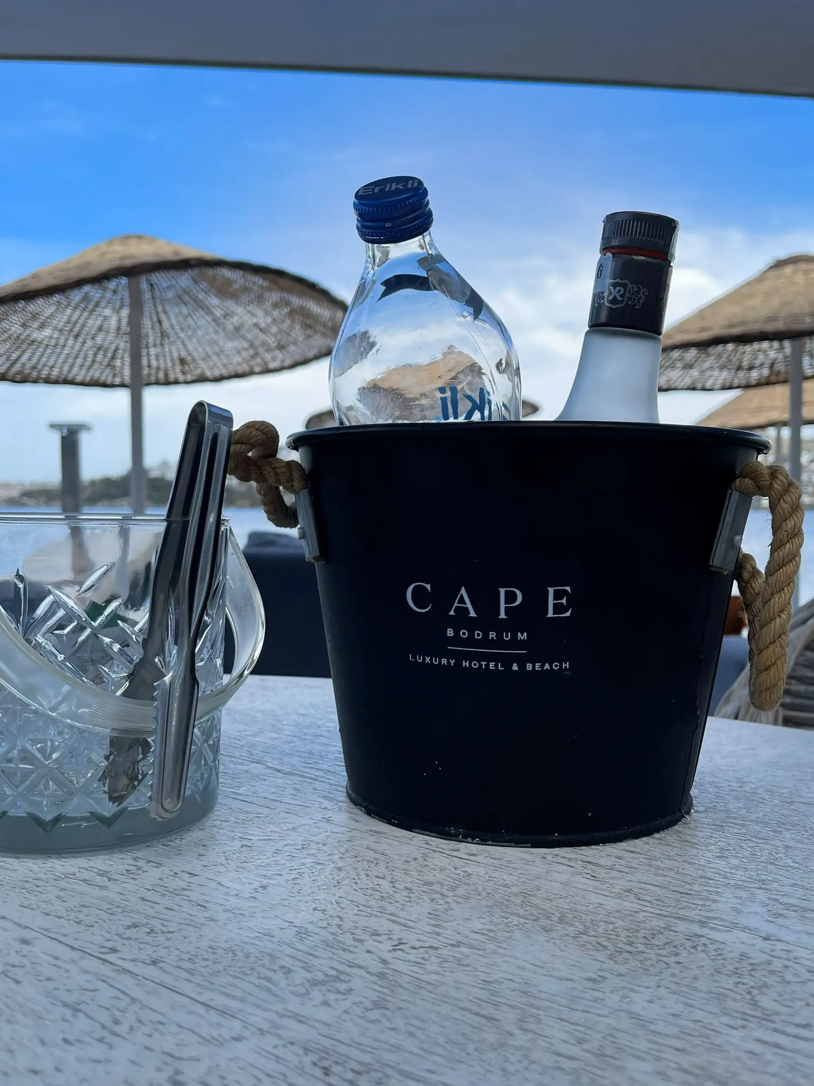 A black ice bucket labeled "CAPE" holds a bottle of clear liquor and a bottle of water on a table. Next to it is a glass with cutlery. Straw parasols and a blue sky are visible in the background.