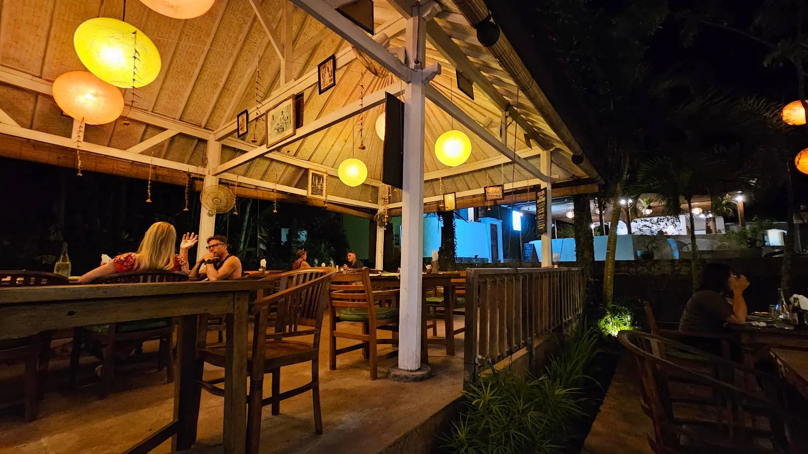 Outdoor restaurant at night with glowing lanterns hanging from a wooden roof. Several people are seated at tables, casually chatting. The warm lighting creates a cozy atmosphere, and there are trees and greenery surrounding the dining area.