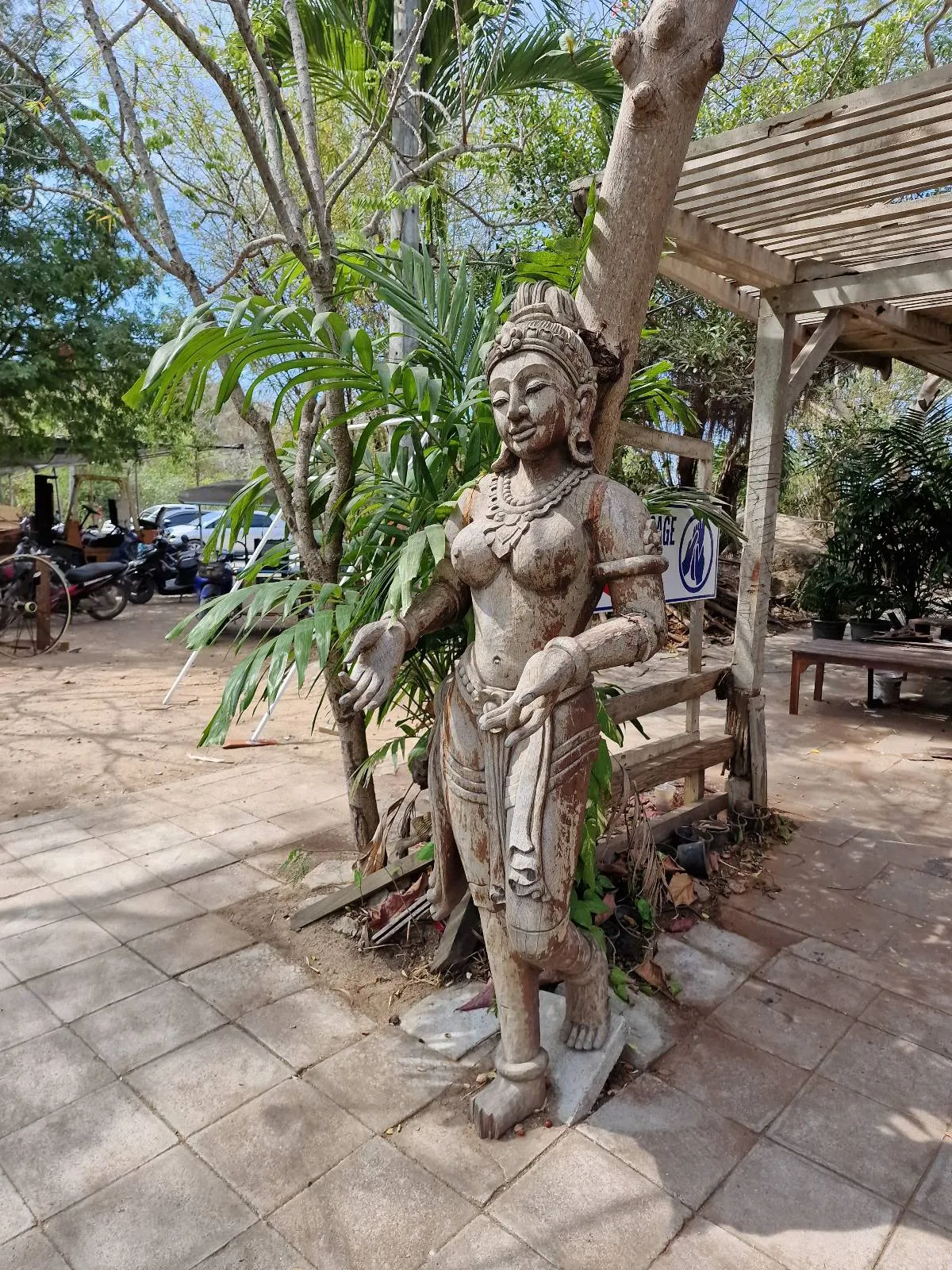 A stone statue of a woman in traditional attire stands outdoors, surrounded by greenery and trees, on a sunlit paved area with benches and a wooden pergola in the background.