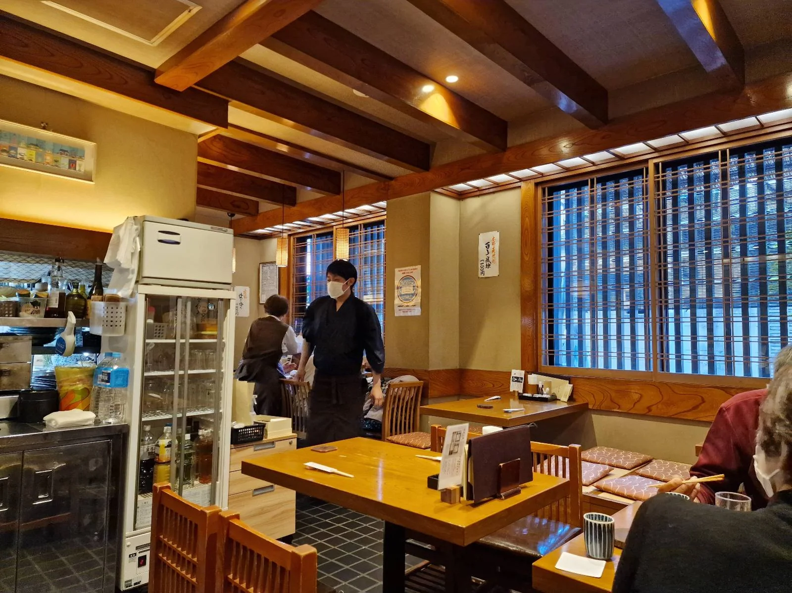 A cozy restaurant interior with wooden beams and warm lighting. There are tables and chairs, a refrigerator near the kitchen area, and a few patrons seated. A staff member in a black uniform is walking through the dining area.