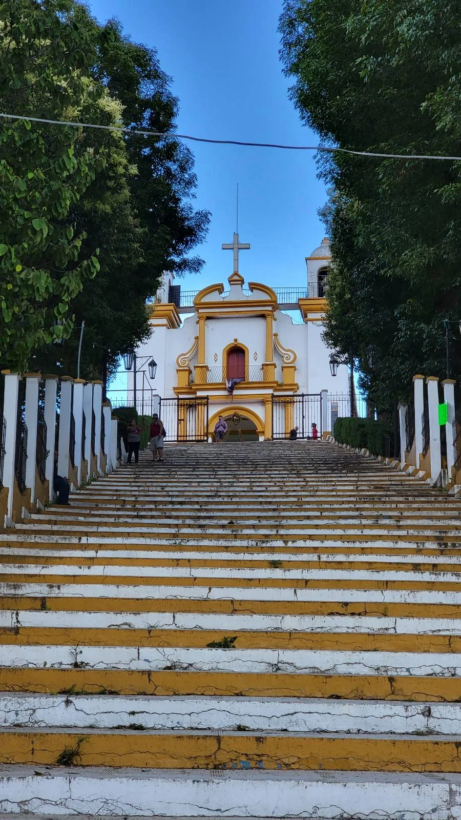 A staircase painted white and yellow leads up to a church with a large cross on its roof. The church has a white facade with yellow accents and is surrounded by trees. The sky above is clear and blue.