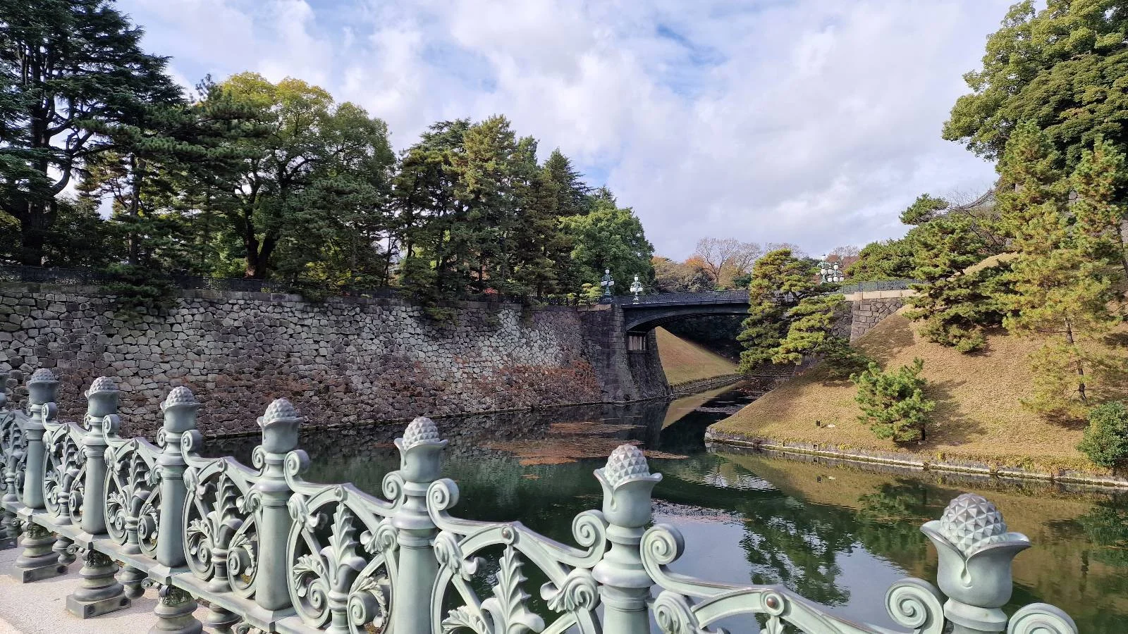 A scenic view of a bridge over a calm river surrounded by lush greenery and a clear sky. The ornate metal railing is visible in the foreground. Trees and stone walls line the riverbank.
