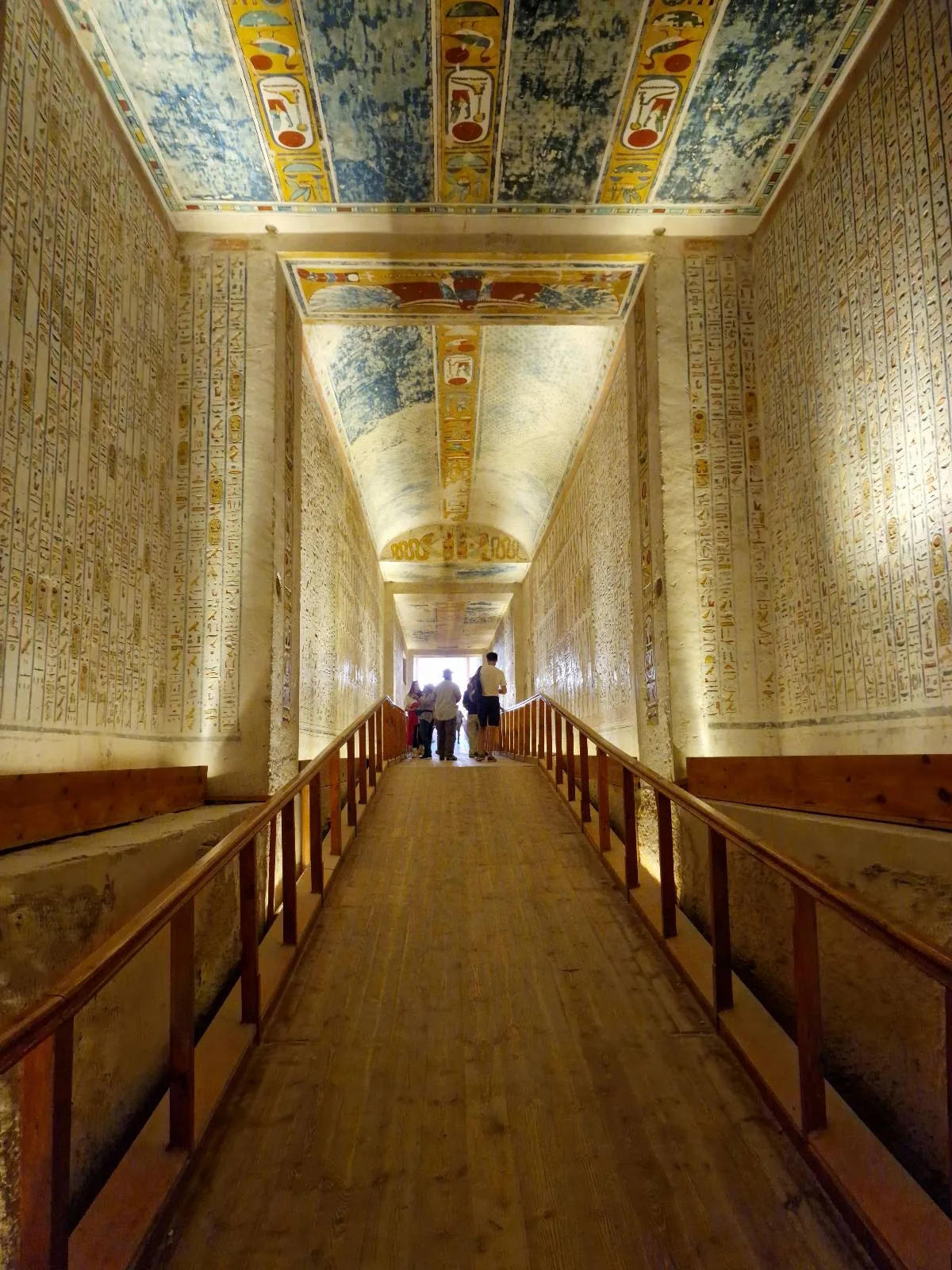 A long, narrow corridor with illuminated beige walls and a wooden walkway. The ceiling features vibrant, intricate artwork. Soft lighting accentuates the details. A few silhouettes of people are visible in the distance.