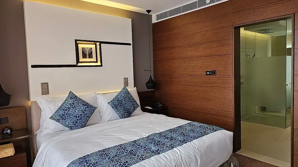 Modern hotel room with a bed featuring blue-patterned pillows and blanket. Wooden wall, framed art, and a frosted glass bathroom door.