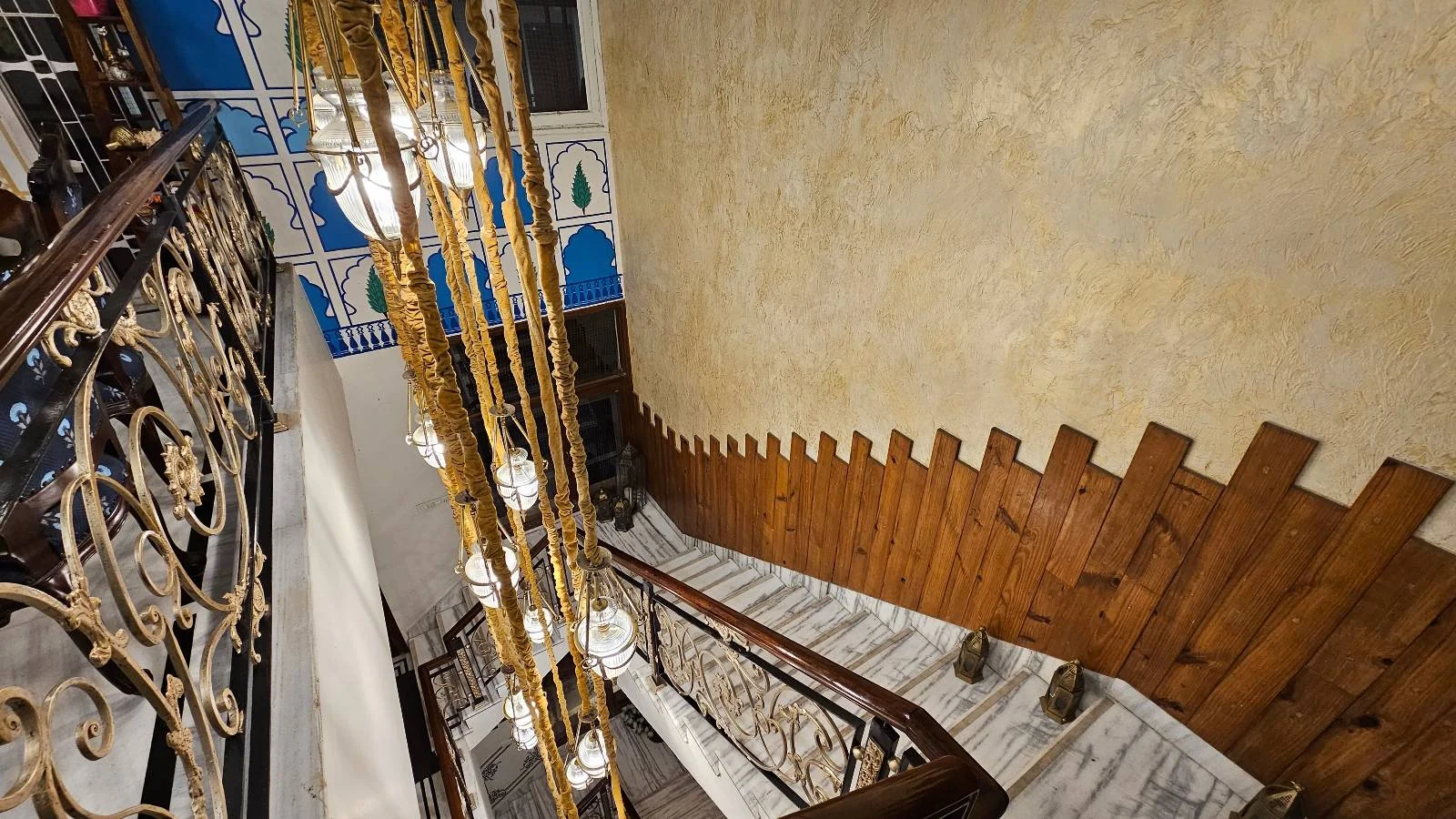 A grand staircase with marble steps, ornate railings, and a gold chandelier with hanging bulbs, against a wall featuring wood accents and decorative blue and white designs.