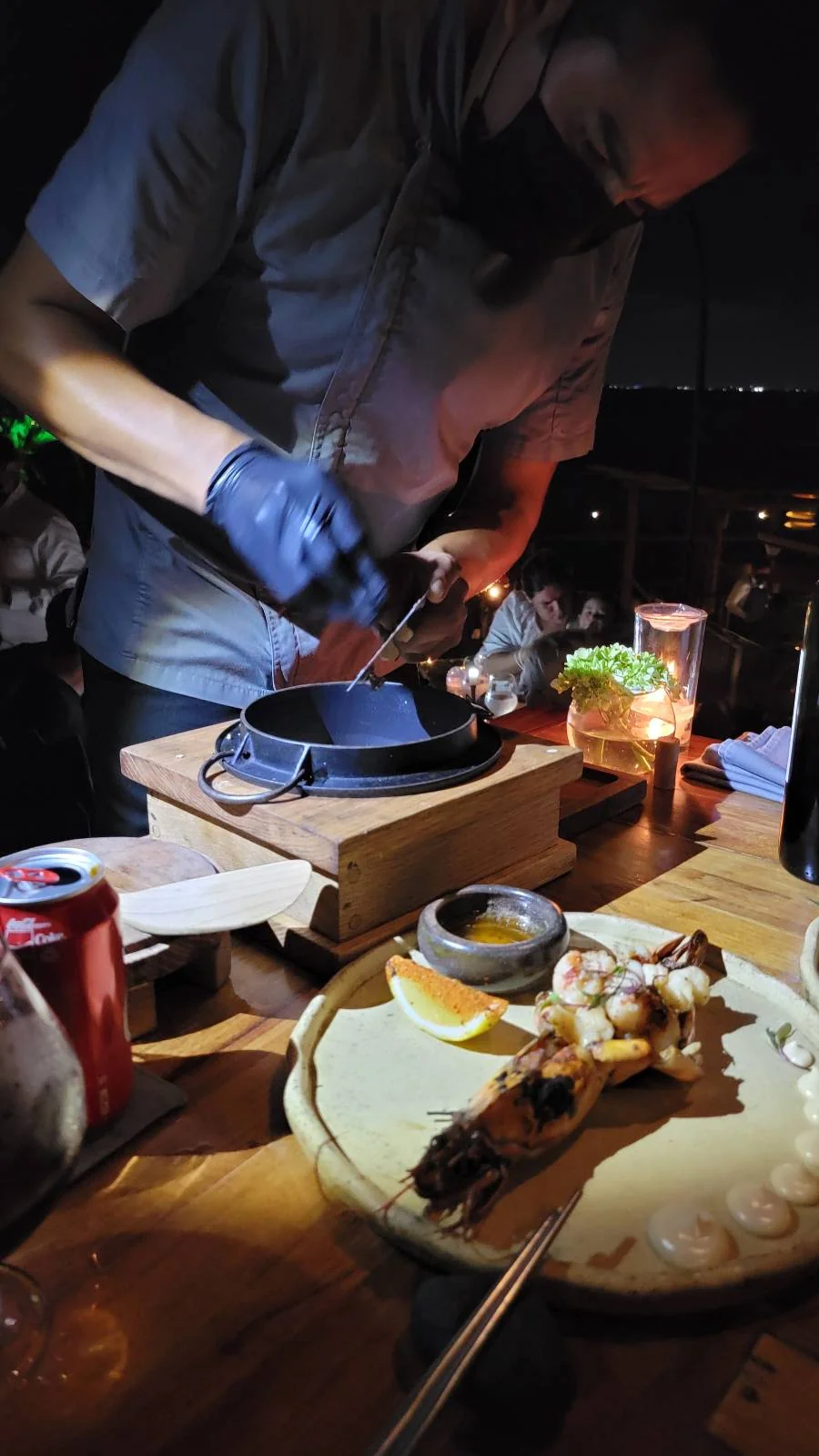 A person wearing a mask and gloves is cooking at a table, using a pan over a small flame. In the foreground, there's a wooden plate with food and a can of soda. The scene is dimly lit with candlelight.