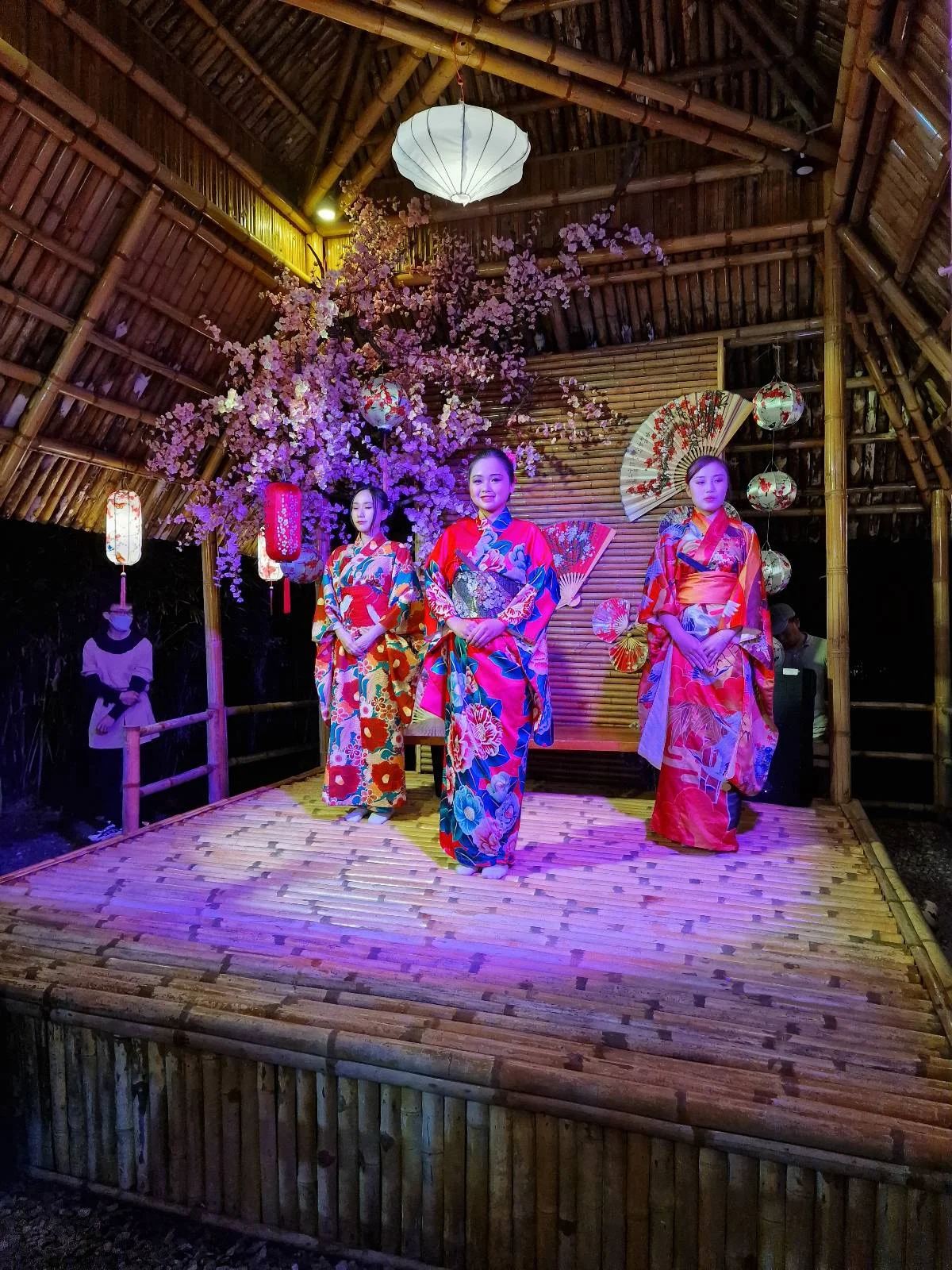 Three women in colorful kimonos standing on a wooden platform adorned with lanterns and cherry blossoms, in a bamboo structure.