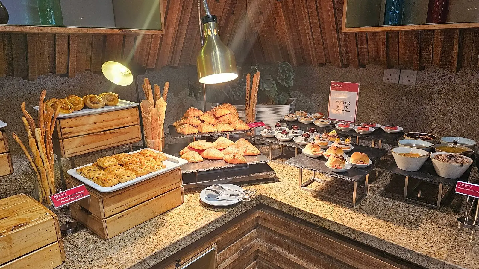 A buffet display featuring an assortment of pastries, including croissants, muffins, and breadsticks, is set on wooden trays and black stands under warm lighting, creating an inviting atmosphere.