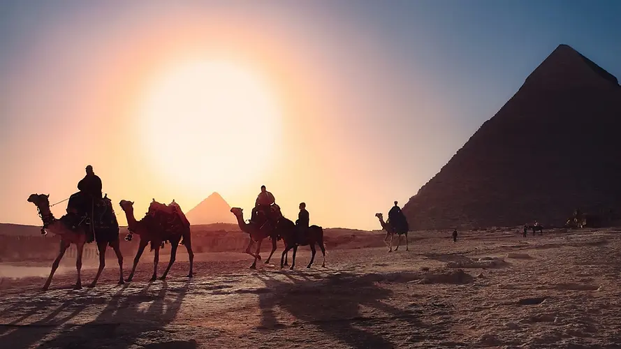 Silhouettes of people riding camels at sunset near the pyramids of Egypt, with the sun low in the sky and large pyramid structures visible in the background.
