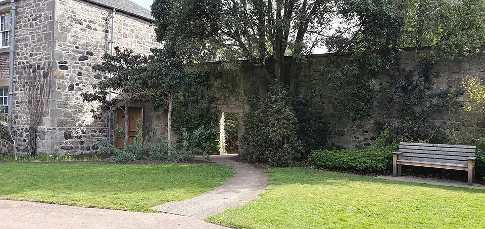 Stone building with arched doorway, surrounded by trees. Path leads to a wooden bench on green lawn, with a serene atmosphere.
