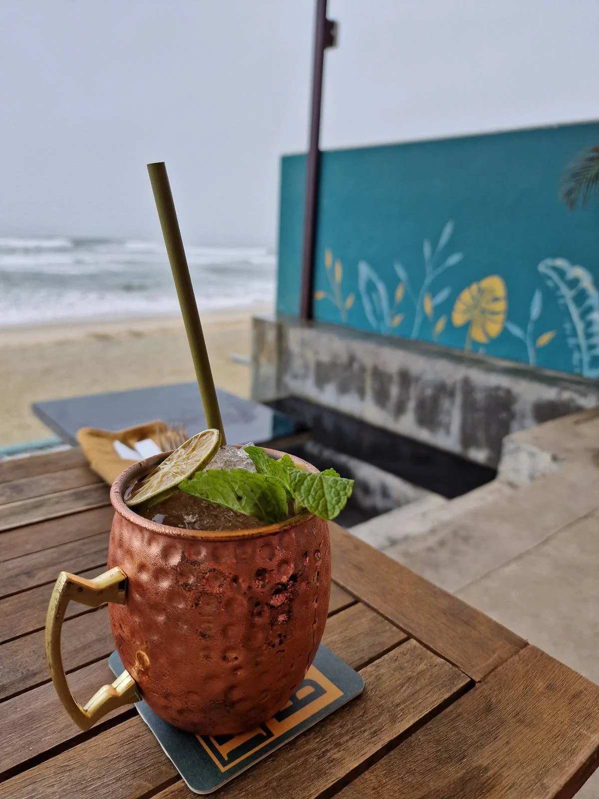A copper mug with a cocktail garnished with mint sits on a wooden table overlooking a beach. The ocean waves are visible in the background, and a teal wall with a floral design is on the side. A straw and coaster complete the setting.