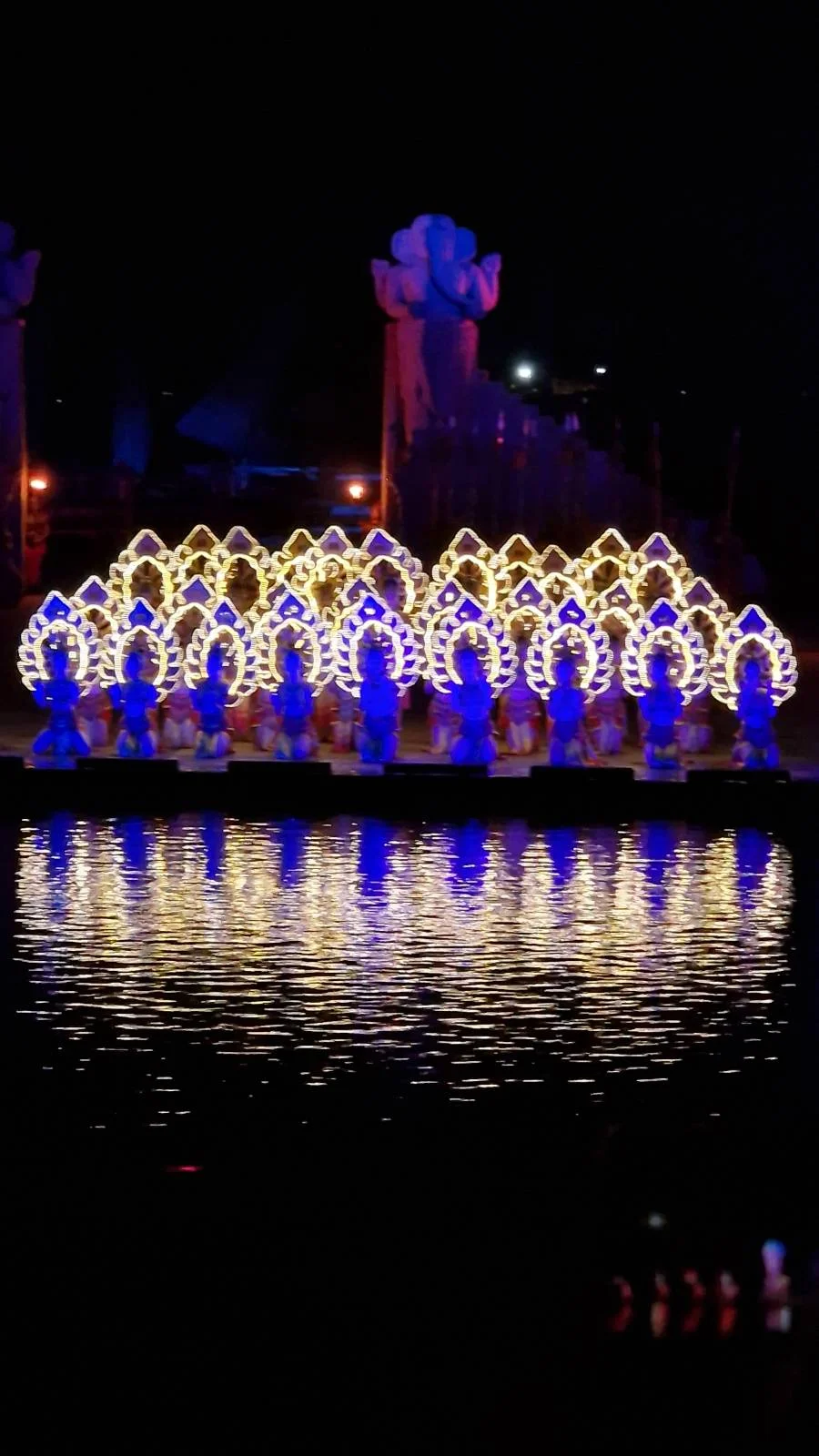 A row of illuminated, ornate figures reflecting on water at night, resembling a traditional Asian design.
