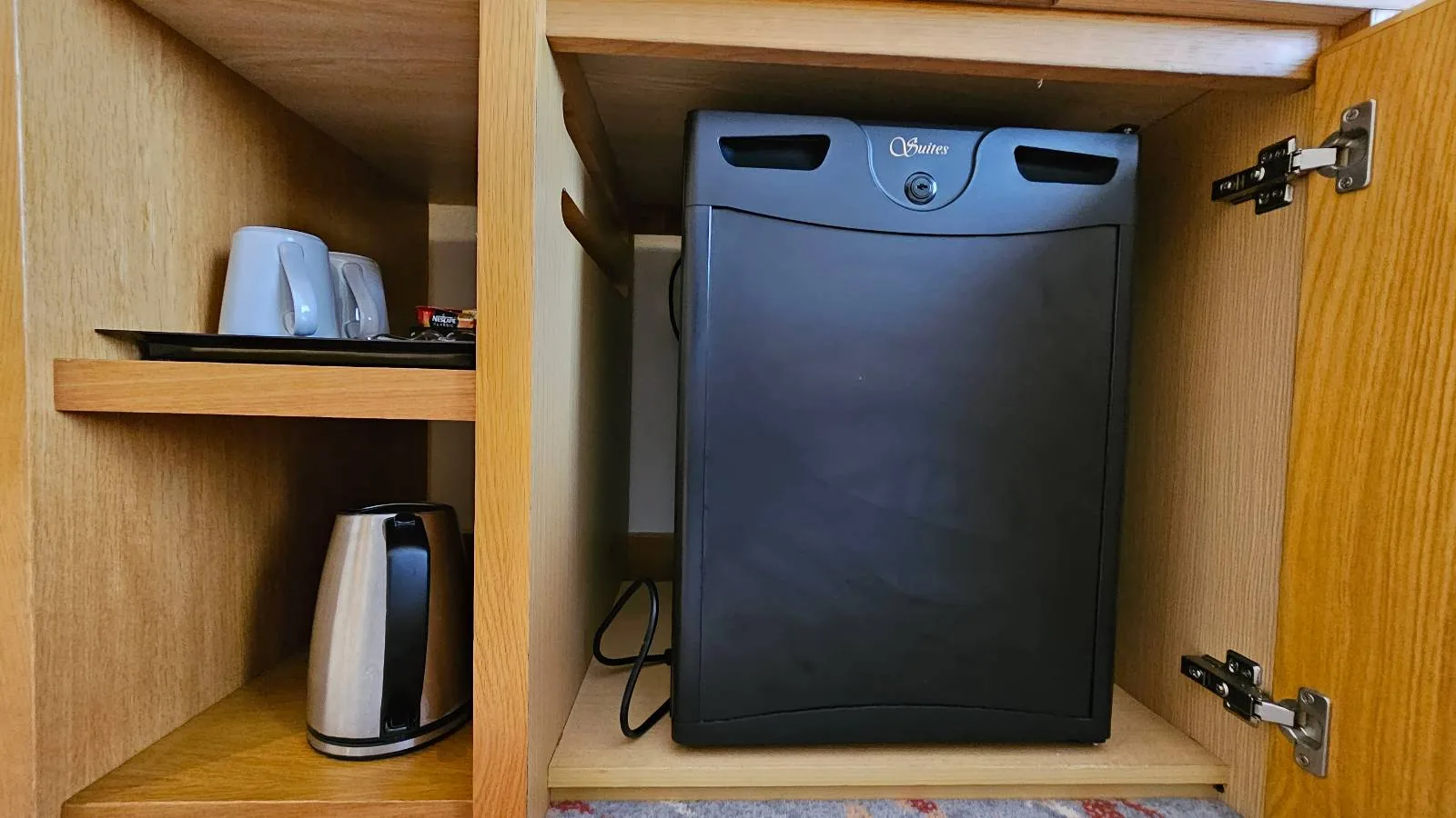A small black refrigerator is placed inside a wooden cabinet. To the left, there is a shelf with a kettle, cups, and other beverages. The cabinet doors are open, revealing the appliances inside.