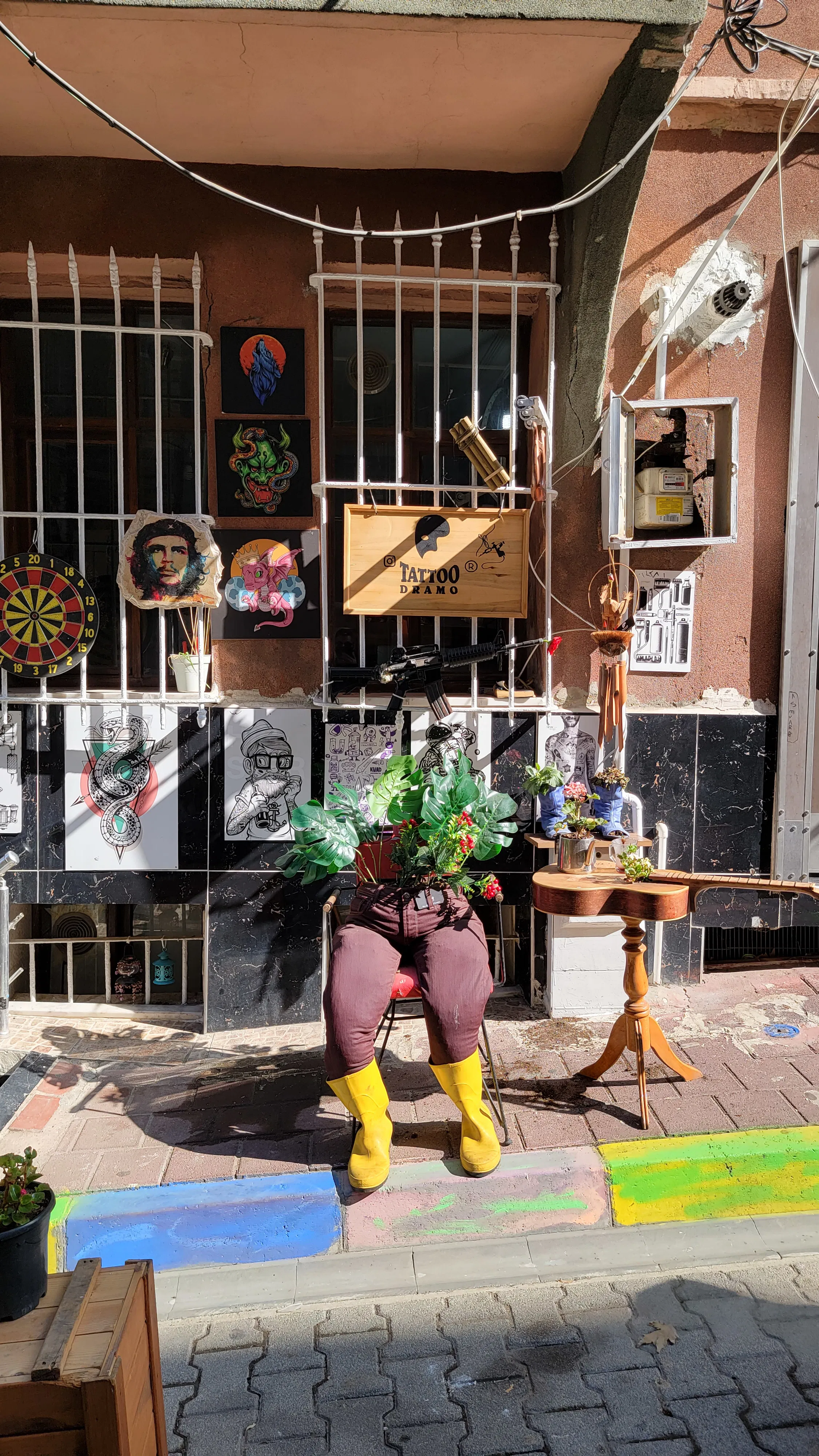A quirky street scene with a mannequin wearing yellow boots and brown pants, holding a plant, sits in front of a building with art pieces, a birdhouse, and a small table against a decorative barred window.
