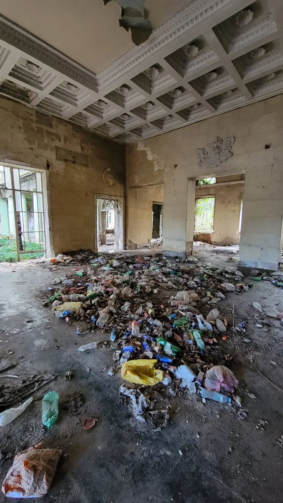A large, abandoned room with high ceilings, dirty walls, and windows without glass. The floor is covered in piles of trash, debris, and discarded items, giving the space a neglected, rundown appearance.
