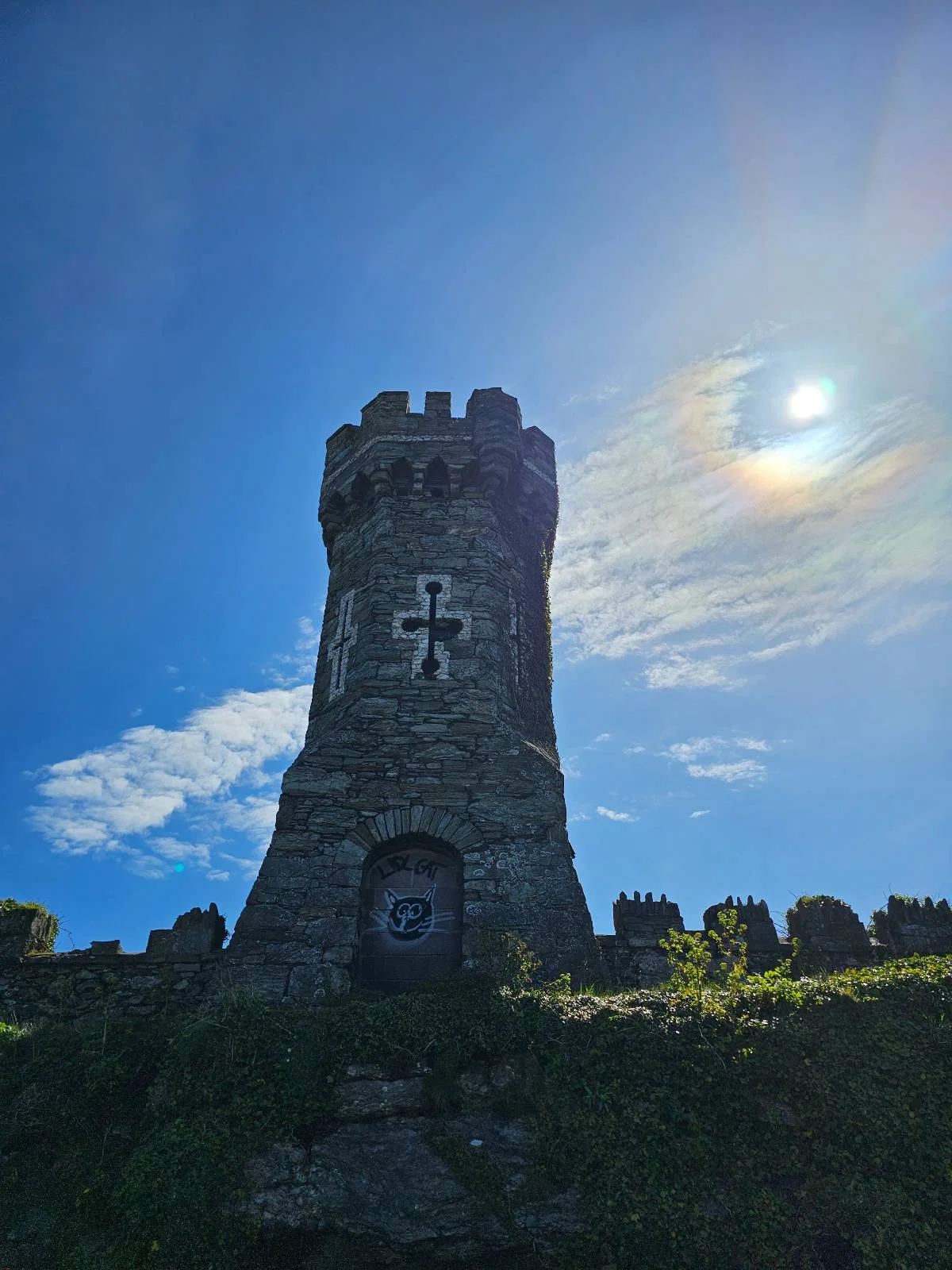 Stone tower with a cross emblem under a clear blue sky, sun partially visible behind the structure.