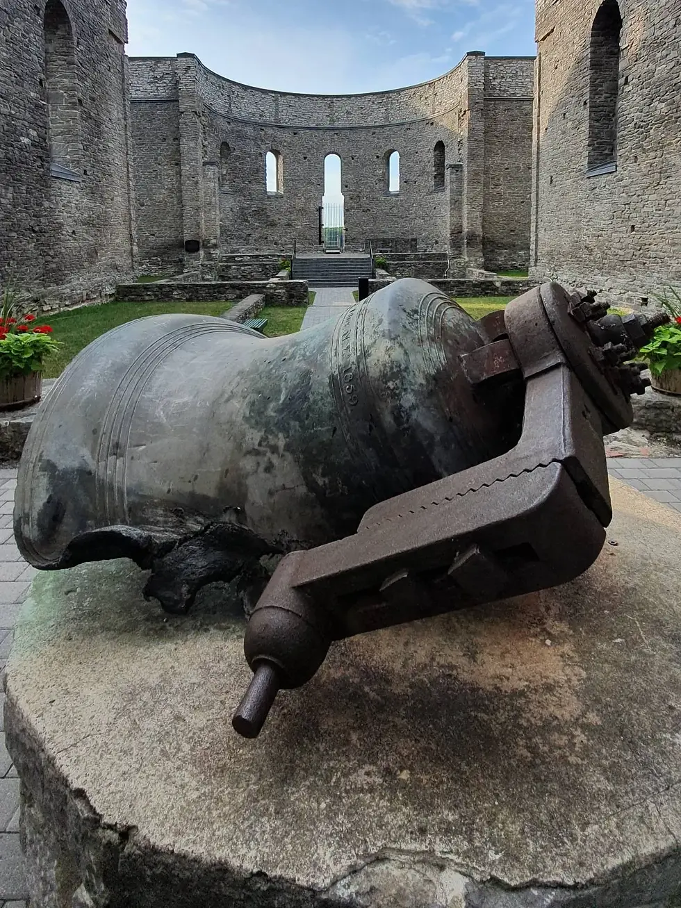 Large cracked bell on stone pedestal in ruins of a stone fortress. Walls with arched windows surround. Sky is clear, mood is historic.