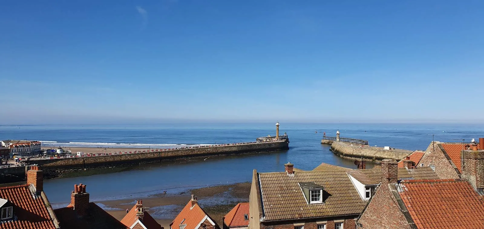 Coastal view with a sandy beach, ocean waves, and a blue sky. Houses and buildings line the left, atop a greenery-covered cliff. A pier extends into the sea, and the tide is low, revealing sand and shallow waters near the shore.