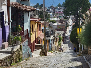 A steep cobblestone street lined with colorful houses and murals meanders through a Mexican village in a lush, hilly area. Power lines and trees are visible against the partly cloudy sky as a person strolls down towards the town centre.
