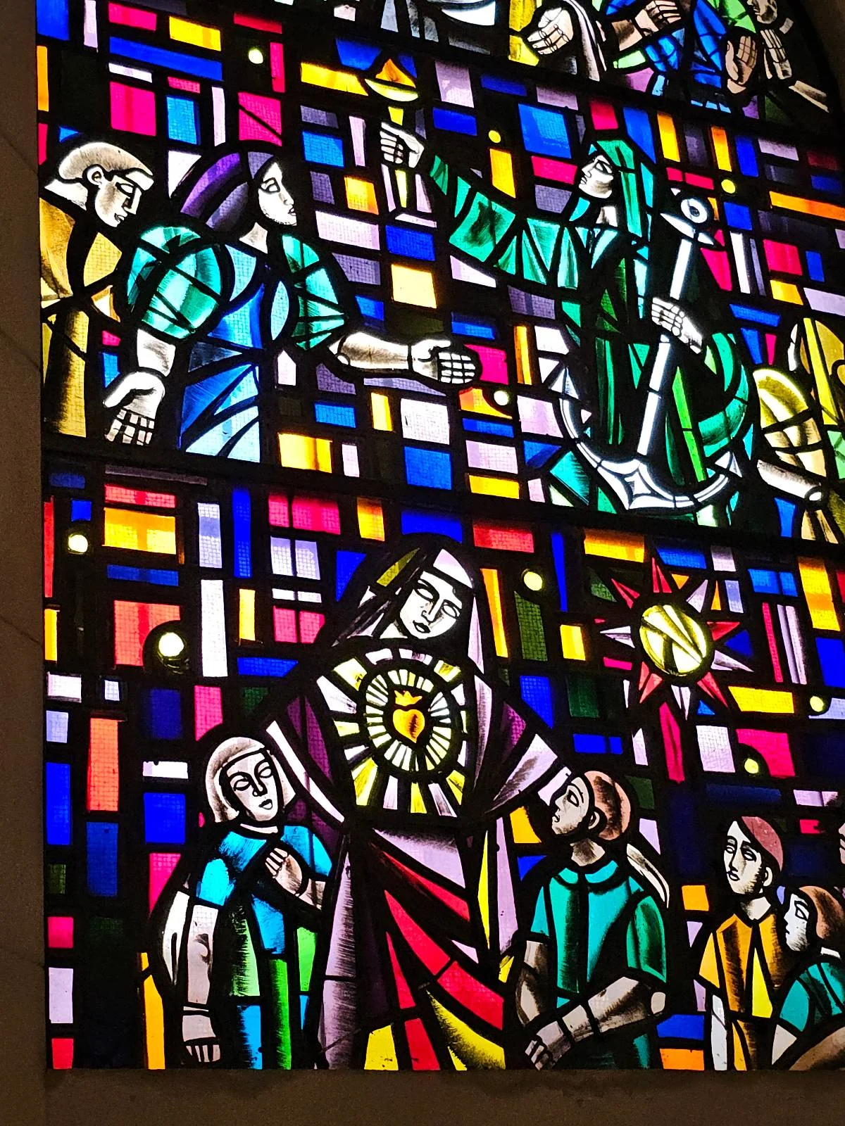 A colorful stained glass window depicts various figures, including a central figure in a robe with a radiant symbol on their chest, surrounded by smaller figures. The background features geometric patterns and vibrant hues.
