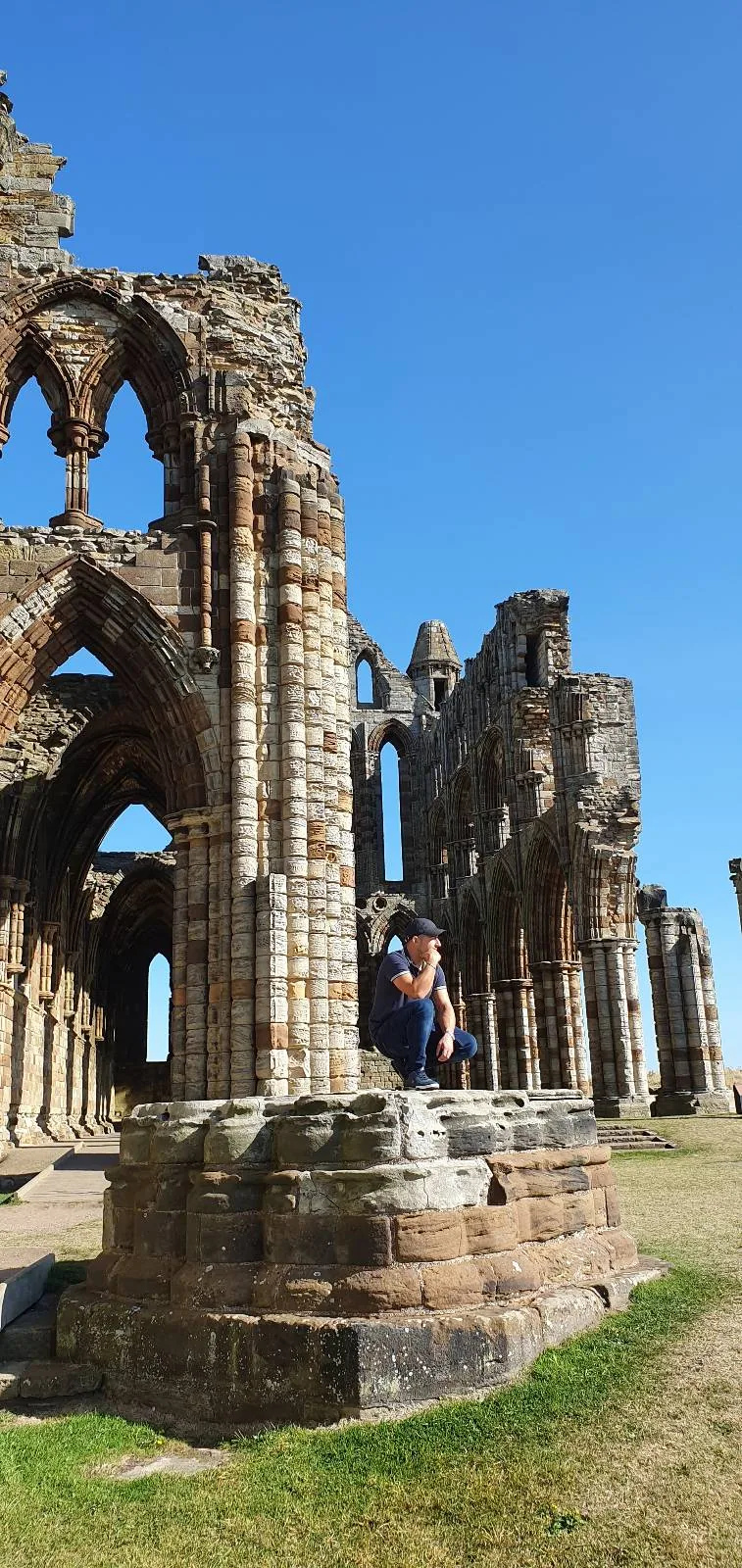A person sitting on a large stone structure, part of ancient stone ruins with tall arches and columns under a clear blue sky. The grass is visible around the ruins, suggesting a historical site or landmark.