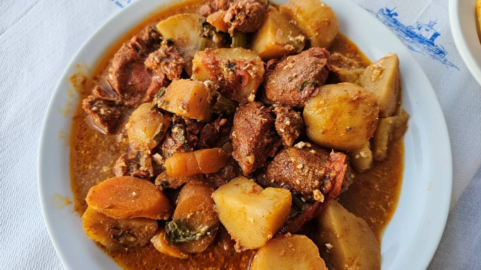 A plate of stew featuring chunks of meat, potatoes, and carrots in a rich, brown sauce, served on a white dish with a blue and white cloth partially visible underneath.