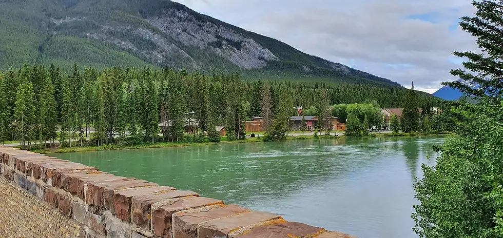 A serene river flows beside a stone wall, bordered by lush green trees and a mountainous backdrop under a cloudy sky.